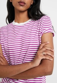 Žena se zkříženýma rukama, oblečená v růžovém a bílém pruhovaném tričku s krátkým rukávem a se zlatým prstenem na levé ruce.