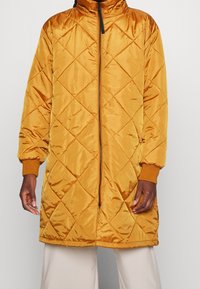 Manteau long matelassé de couleur jaune moutarde, fabriqué en tissu brillant. Il comporte un col montant, une fermeture éclair devant et des poignets côtelés. Semble léger.