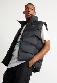 Sort puffer vest med høj krave, lynlås lukning og polstret design. Båret over en hvid T-shirt med blå grafisk detalje og sorte bukser.