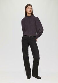 Dunkellila, langärmlige Bluse mit rüschendem Ausschnitt und glitzerndem Detail, kombiniert mit schwarzen Schlagjeans und Ankle Boots.