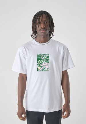 Mann mit Dreadlocks, der ein weißes T-Shirt mit einer grünen Grafik und dem Schriftzug "CLEPTOMANICX" trägt, steht vor einem schlichten Hintergrund.