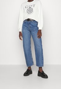 Felpa bianca con logo GANT nero, jeans blu a gamba larga con cuciture visibili e mocassini neri con zeppa. Design semplice e vestibilità casual.