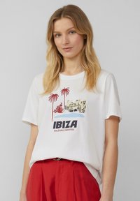 T-shirt blanc avec un graphique coloré de palmiers et une scène avec le texte "IBIZA" et "PARADIS BALEARES". Manches courtes, coupe décontractée.