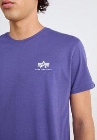 Vijolična bombažna majica s kratkimi rokavi in krog oblikovanim ovratnikom, z belim logotipom na levem prsnem delu. Gladka tekstura, priložnosten kroj.