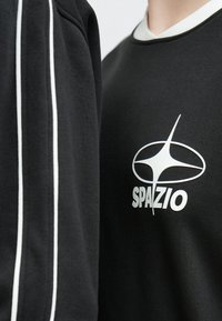 Fekete pulóver fehér "SPAZIO" felirattal és vékony fehér csíkokkal az ujjakon. Puha, sima tapintású anyagból.