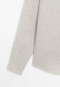 Poignet de manche longue et partie inférieure latérale d'une chemise en lin beige avec texture visible du tissu sur fond blanc.
