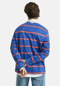 Blå langermet skjorte med oransje horisontale striper, hvit krage, avslappet passform og sideslitser i nederkant. Modellen står med hendene bak ryggen.