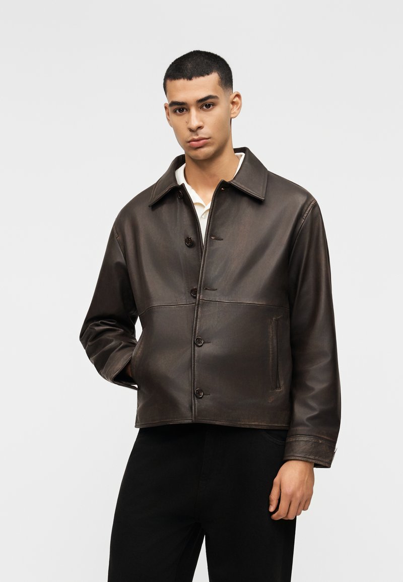 Νεαρός άνδρας με κοντά μαλλιά, φοράει ένα σκούρο καφέ δερμάτινο μπουφάν με κουμπιά πάνω από ένα λευκό πουκάμισο, στέκεται με το ένα χέρι στην τσέπη.