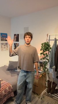 Noormees vabaaja riietes tõstab paremat kätt magamistoas, kus on voodi, riputatavad riided ja rohelise võraga taim kapil.