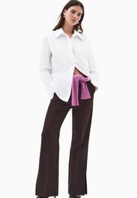 Hvid skjorte med knapper og lomme, parret med brune flagrende bukser. En lyserød sweater bundet om taljen tilføjer farvekontrast.