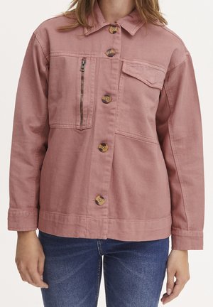 Kvinde iført en støvet lyserød jakke med knapper og en lodret lynlåslomme samt blå jeans, stående mod en hvid baggrund.