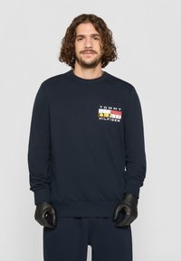 Mørkeblå hettegenser med ribbet rund hals og lange ermer. Har en fargerik logo på øverste venstre bryst. Myk tekstur i stoffet.