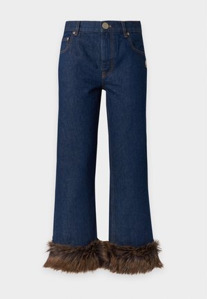 Femme portant un haut blanc sans manches et un jean en denim foncé avec des poignets en fourrure marron, debout avec une main dans la poche et des talons noirs.