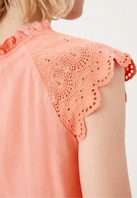 Blusa pescozza con dettagli in pizzo ricamato sulle spalle, con bordo scallop e una consistenza morbida e liscia sul corpo.