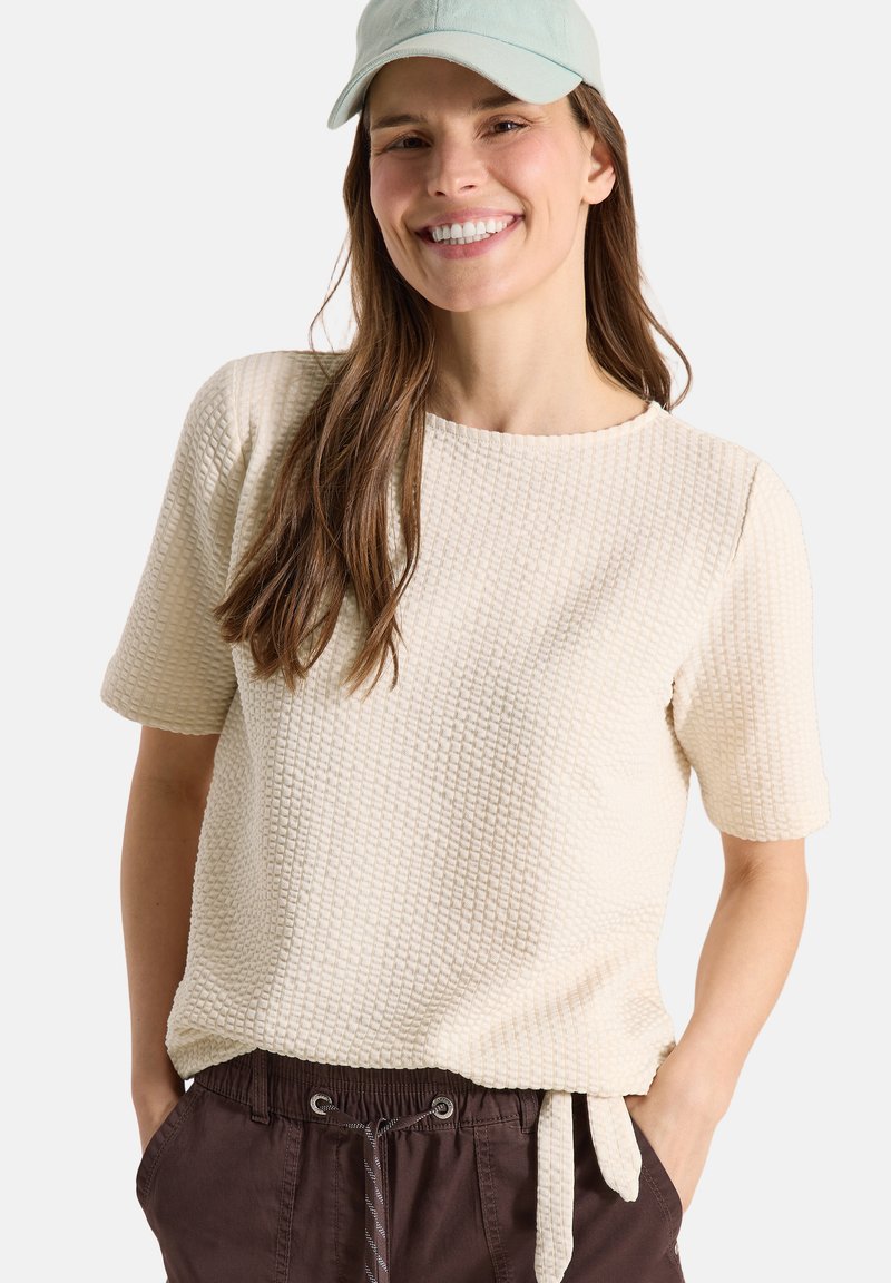 Femme souriante portant une casquette bleu clair, un haut texturé crème à manches courtes et un pantalon marron foncé à cordon, les mains dans les poches, sur fond blanc.