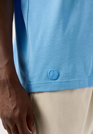 Dark-skinned hand rests beside light blue short-sleeve shirt with small round JOTT logo above beige pants.