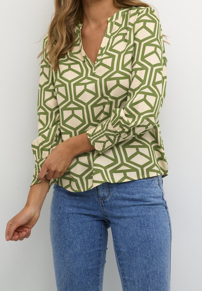 Femme portant un chemisier crème avec des motifs géométriques verts et un jean bleu, ajustant le poignet de sa manche longue.