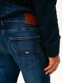 Jeans de mezclilla azul oscuro con un acabado texturizado, que presentan un parche de cuero marrón etiquetado "TOMMY JEANS" y detalles de costura visibles.