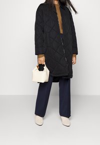 Žena v černém prošívaném kabátu, hnědém topu, námořnických kalhotách s širokými nohavicemi, krémových pletených kotníčkových botách, držící malou krémovou kabelku.