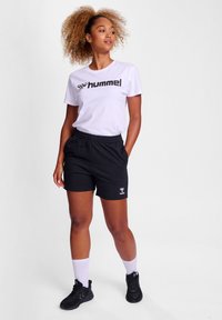 Hvid t-shirt med sort "hummel" logo, parret med sorte shorts, hvide sokker og sorte sportssko. Modellen står med hænderne på hoften.