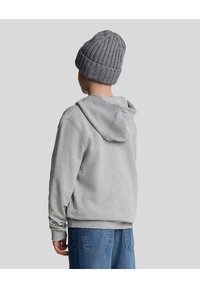 Szary, kapturowy sweter o miękkiej fakturze, z kieszenią kangurką. W połączeniu z dzianinową, prążkowaną, szaro-beżową czapką oraz niebieskimi dżinsami.
