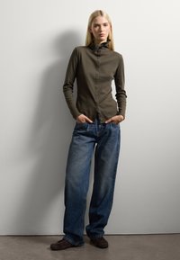 Junge Frau mit langen blonden Haaren, die einen knöpfbaren olivgrünen Rollkragenpullover, lockere blaue Jeans und dunkle Schuhe trägt, steht mit den Händen in den Taschen.