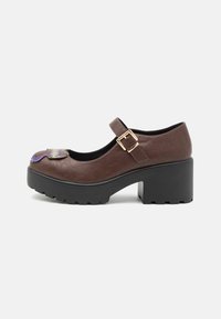 Brązowe buty typu Mary Jane z teksturowanym syntetycznym wierzchem, masywnym czarnym obcasem, detalem z klamrą i dekoracyjnym akcentem w okolicy palców.