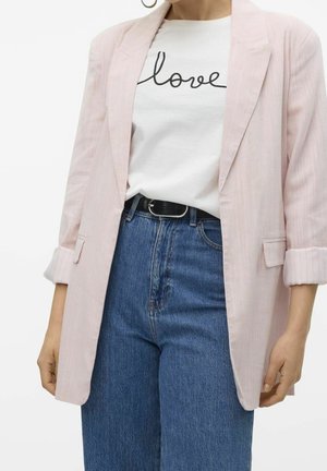 Femme portant un blazer rose pâle, un t-shirt blanc avec le texte "love", un jean taille haute bleu et une ceinture noire, montrée de l'épaule aux hanches.