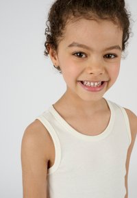 Top in cotone chiaro, senza maniche, con scollatura rotonda e spalline larghe; modella sorridente con capelli ricci e orecchini a cerchio.