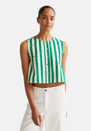 Mujer con top corto sin mangas a rayas verticales verdes y blancas abotonado, con pantalones blancos, de pie frente a un fondo liso.