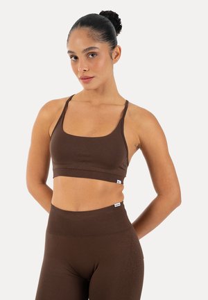 Vrouw met donker haar in een knot, gekleed in een bruine mouwloze crop top en hooggetailleerde legging tegen een egale lichte achtergrond.