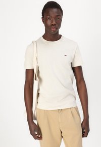Lys beige bomull t-skjorte med rund hals og liten logo-detalj. Kombinert med lyse brune bukser og en kremfarget tote-bag.