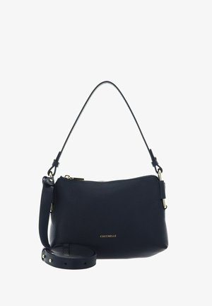 Sac à main Coccinelle en cuir noir avec fermeture éclair sur le dessus, bandoulière réglable et poignée courte sur fond blanc.