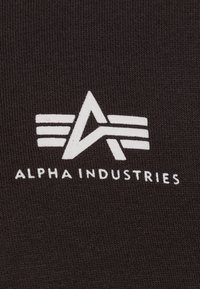 Černá tkanina s bílým logem s úhlovým designem a horizontálními pruhy, pod logem je nápis "ALPHA INDUSTRIES".