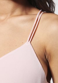 Close-up van de schouder en bovenborst van een vrouw die een lichtroze mouwloos topje draagt met dunne dubbele banden.