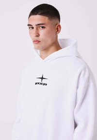 Sudadera blanca con capucha, hecha de un material suave, que presenta un logo negro "PXP" y un gráfico de estrella en el pecho, con un ajuste relajado y una capucha grande.