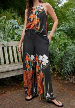 Combinaison noire avec des motifs floraux vibrants orange et blancs, coupe ample, sans manches et taille élastiquée. Associée avec des tongs.