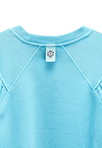 Jasnoniebieska bluza z dekoltem w serek, wykończona prążkowanym kołnierzem i małą metką z tyłu z szarym kwiatem o czerwonym środku.