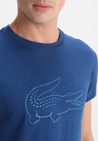 Blå bomulls t-shirt med en texturerad krokodillogo i en ljusare blå nyans. Rundhalsdesign med korta ärmar. Slät textur.