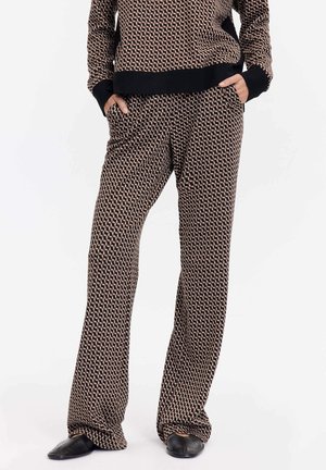 Personne portant un ensemble assorti à motifs géométriques : haut à manches longues et pantalon large avec bordure noire, mains dans les poches, chaussures noires, fond blanc.