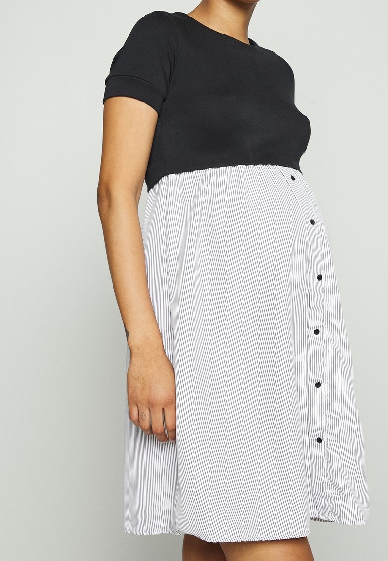 Femme portant une robe noire à manches courtes avec une jupe blanche à rayures verticales et des boutons noirs, debout devant un fond uni.