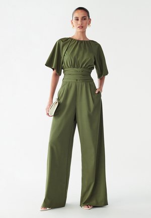 Olivengrøn buksedragt med vide ben, løse korte ærmer, rynket halsudskæring og tilspidset talje. Modellen holder en lille cremefarvet håndtaske.