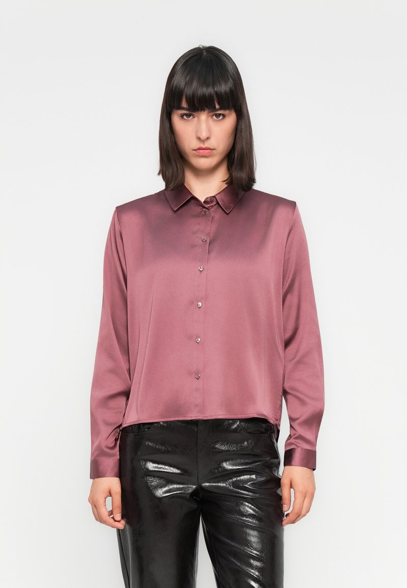 Camisa de manga longa, abotoada, em cetim de mauve profundo. Design com colarinho e um corte ligeiramente relaxado, com um brilho subtil, combinada com calças pretas brilhantes.