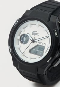 Μαύρο ψηφιακό ρολόι με στρογγυλό άσπρο καντράν, που διαθέτει μικρή ψηφιακή οθόνη, λευκούς δείκτες και μαύρο λαστιχένιο λουράκι με υφή.