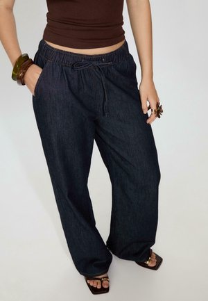 Person wearing dark blue wide-leg denim pants with elastic waist and drawstring, brown sleeveless top, chunky bracelets, rings, and open-toe sandals.