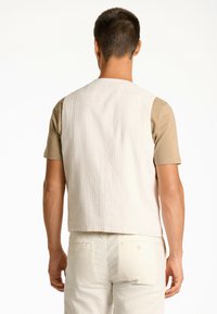 Beige Baumwollweste mit einem schlichten Design, das eine strukturierte Oberfläche, einen runden Halsausschnitt und einen geraden Schnitt aufweist. Wird über einem hellbraunen Hemd getragen.