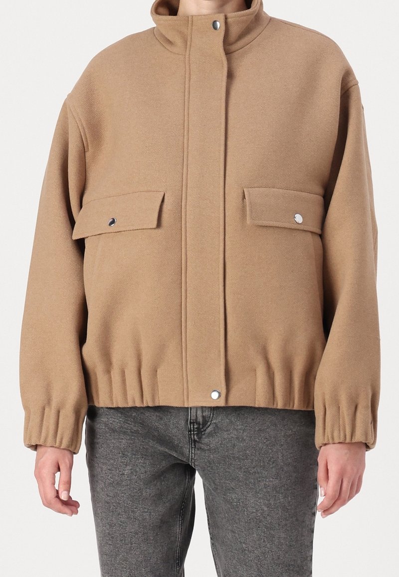 Giacca marrone oversize con collo alto, chiusura a bottoni a pressione e due tasche frontali; orlo a costine e tessuto strutturato.