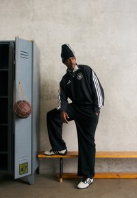 Svart Adidas treningsdrakt med tre hvite striper, stort logo på brystet, iført en svart lue, sittende ved siden av et blått skap og en basketball.