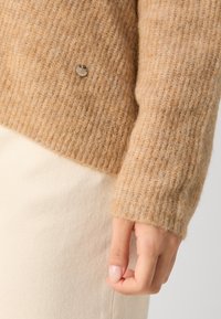 Manche de pull en maille beige duveteuse avec poignet côtelé, petite étiquette ronde en métal de la marque, et main reposant contre un tissu crème clair.