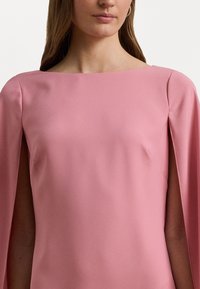 Vaaleanpunainen, kevyt mekko, jossa on neliömäinen pääntie ja viittahuput. Sileä pinta, minimalistinen muotoilu, eikä printtejä tai koristeita.